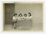 Four women working at desk in front of barrack
